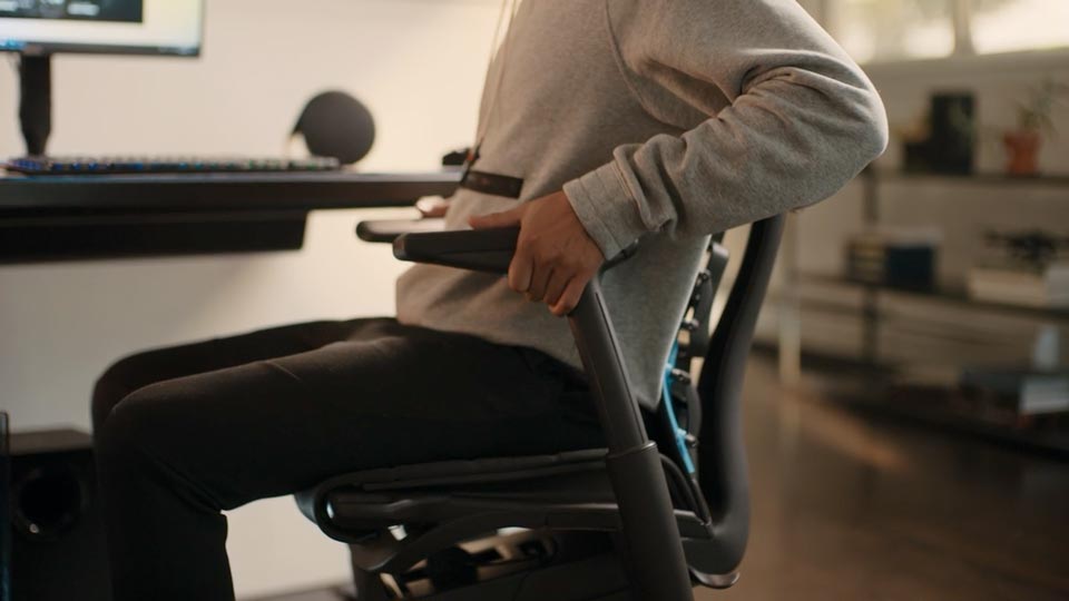 En video av en person som sitter i en svart och blå Embody-spelstol från Herman Miller och sträcker sig bredvid stolen för att justera armstöden.