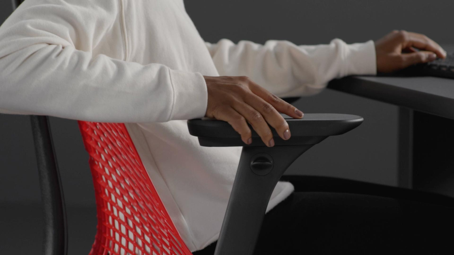 En video av en person som sitter i en röd och svart Sayl-gamingstol från Herman Miller och sträcker sig bredvid stolen för att justera armstöden.