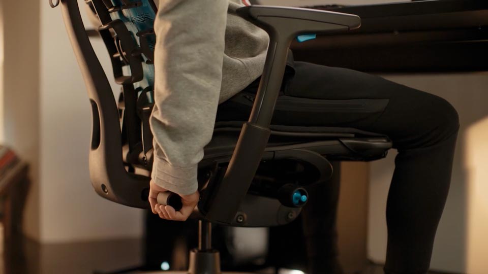 En video av en person som sitter i en svart och blå Embody-spelstol från Herman Miller och sträcker sig bakom stolen för att justera ryggstödet.