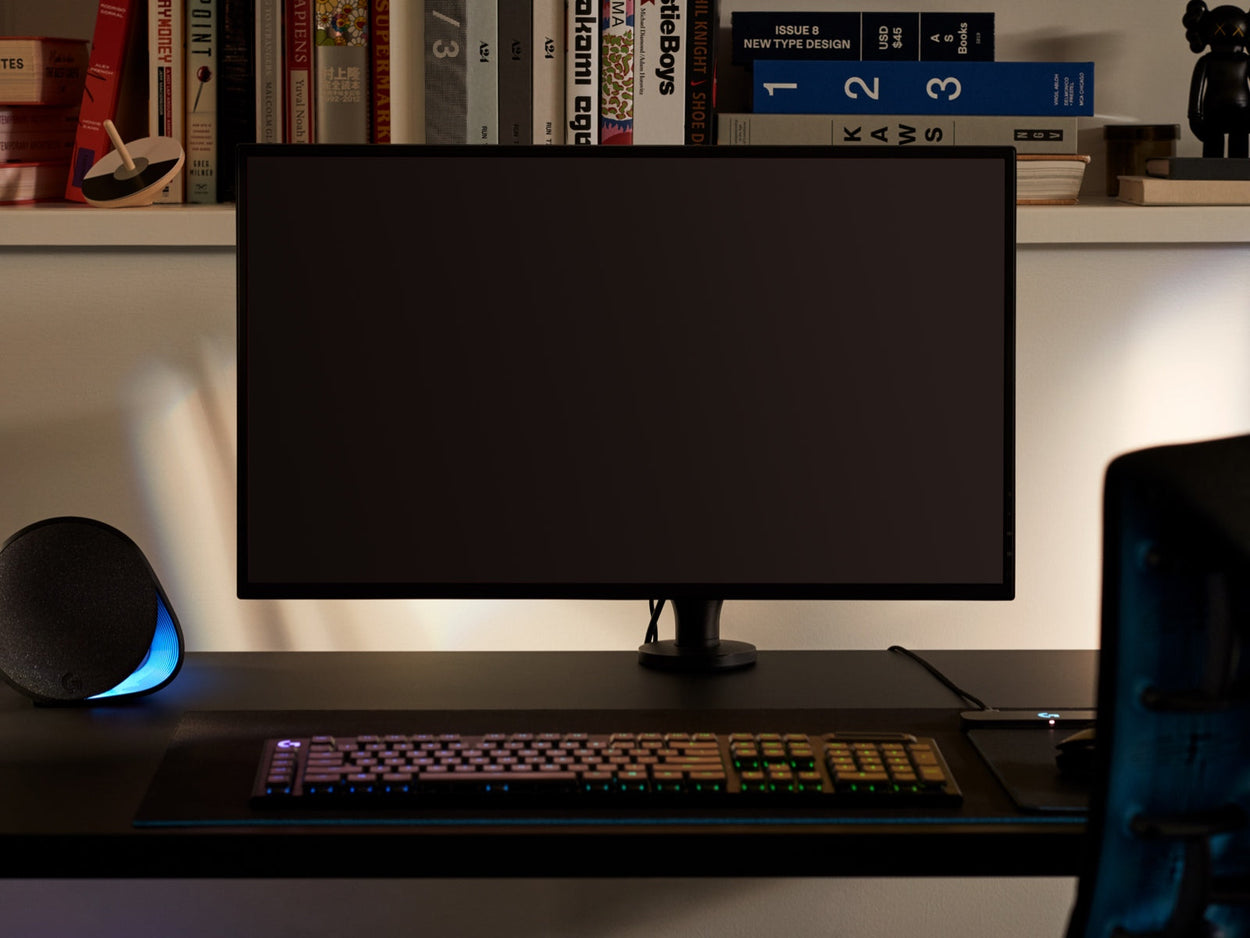 Framifrån sett av en svart Ollin-skärmarmshållare som håller en datorskärm och är fäst vid ett svart Nevi-spelskrivbord från Herman Miller. Placerat i ett spelrum eller hemmakontor, med en bokhylla, tangentbord och mus.