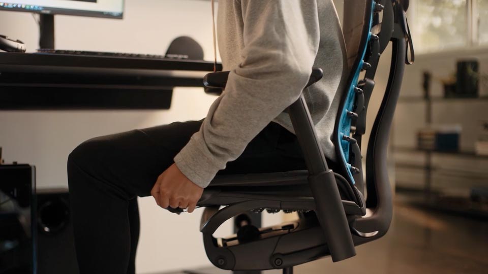 En video av en person som sitter i en svart och blå Embody-spelstol från Herman Miller och sträcker sig bredvid stolen för att justera sittdjupet.
