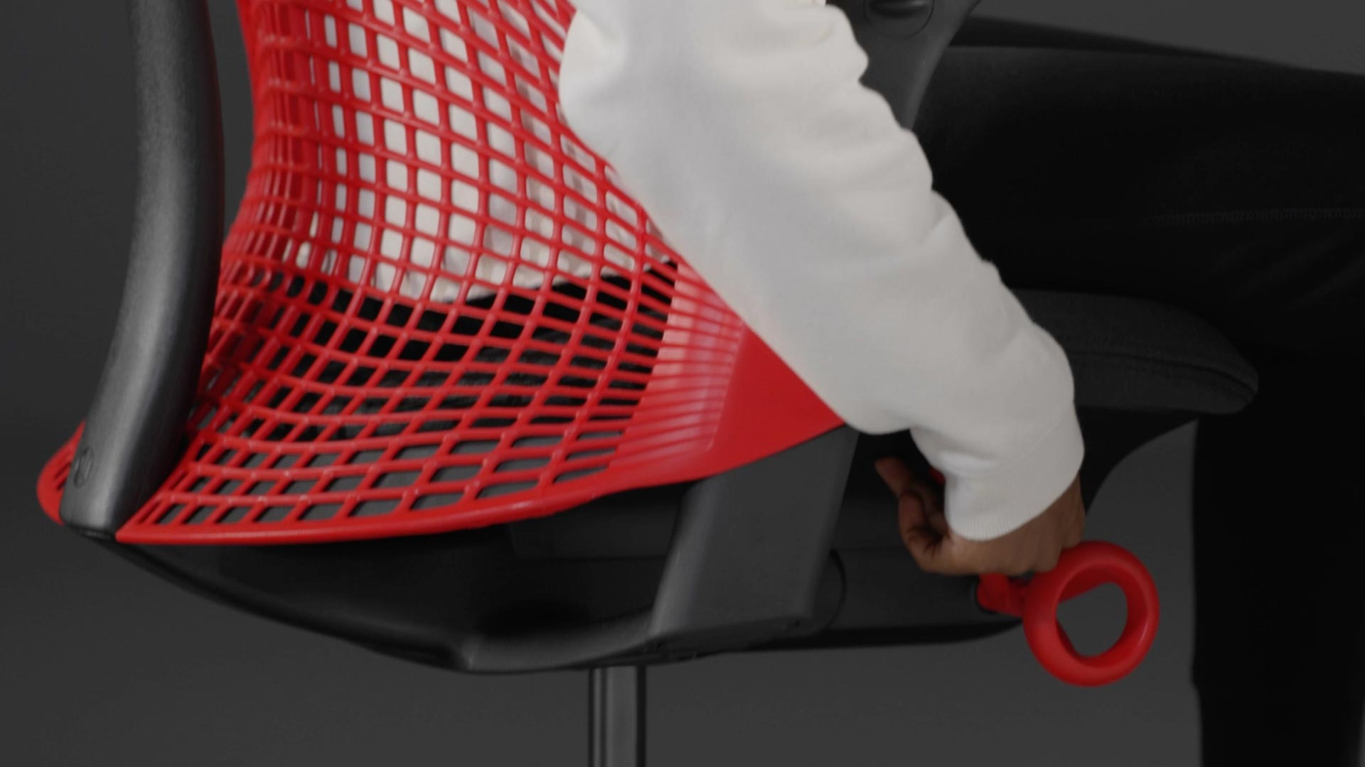 En video av en person som sitter i en röd och svart Sayl-gamingstol från Herman Miller och sträcker sig bredvid stolen för att justera sitthöjden.