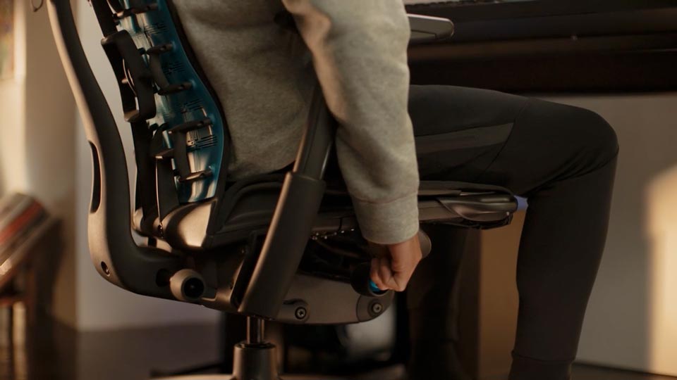 En video av en person som sitter i en svart och blå Embody-spelstol från Herman Miller och sträcker sig bredvid stolen för att justera sitthöjden.