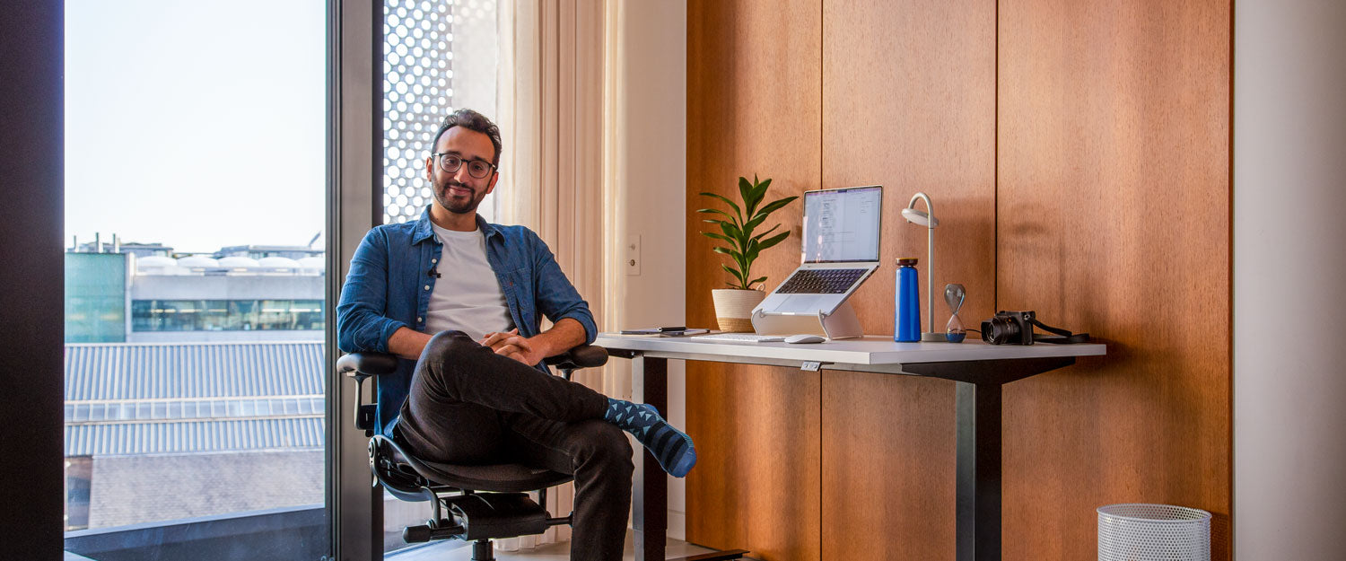 Foto av Ali Abdaal, poddare, sittande i Herman Miller Aeron kontorsstol i grafit framför Nevi sit-stand-skrivbord med svarta ben och vit skiva med en bärbar dator i ett Oripura-laptopstativ
