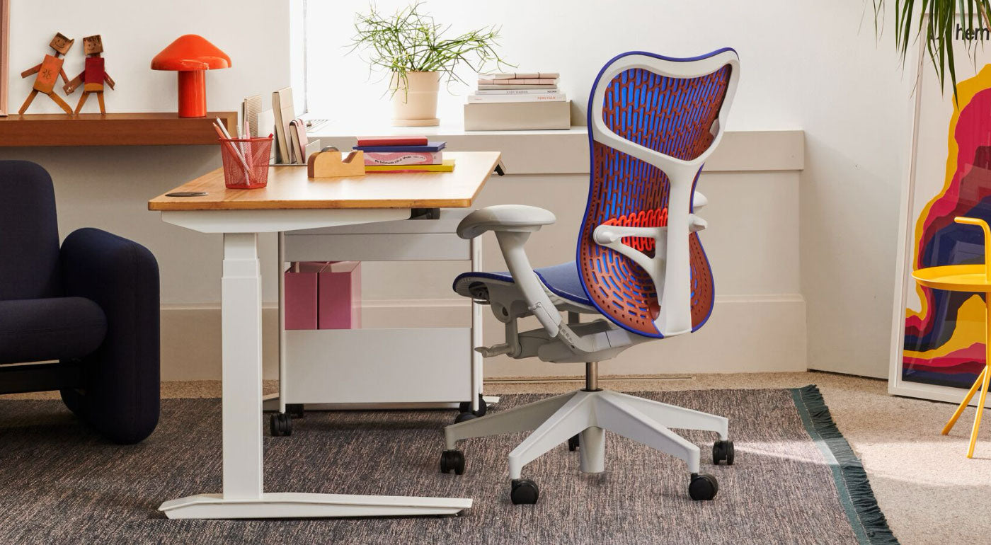 En blå, orange och vit Mirra 2-kontorsstol från Herman Miller, placerad bredvid ett Jarvis-ståbord. Placerad i en hemmakontorsmiljö med en blå Wilkes-soffa, blå Maharam-matta, gult DLM-sidobord och röd HAY Pao-bordslampa.