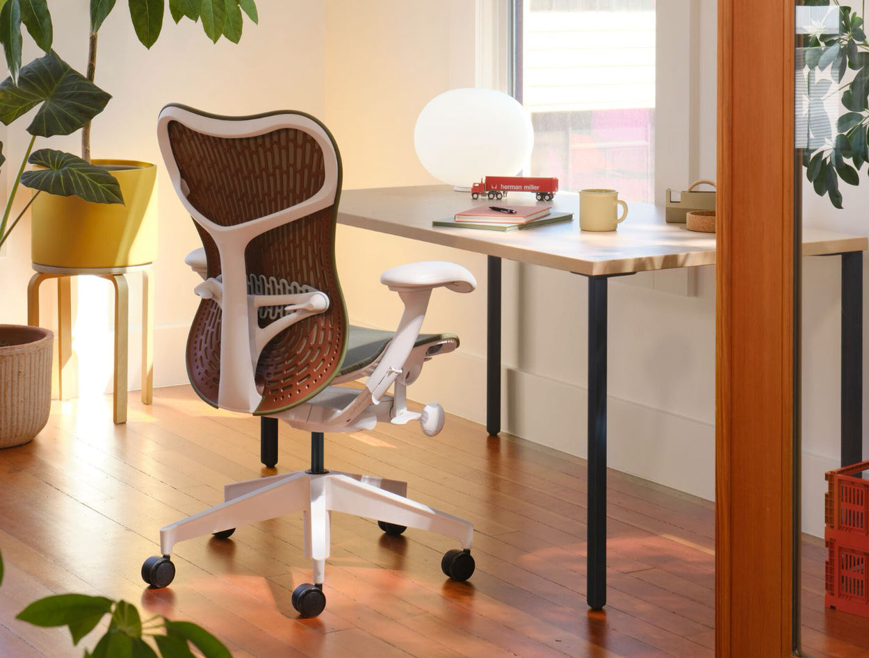 En olivgrön och vit Mirra 2-kontorsstol med rödbrunt ryggstöd, placerad framför ett Herman Miller OE1-skrivbord. Placerad i en hemmakontorsmiljö med massivt trägolv, vita väggar och varm, mysig belysning.