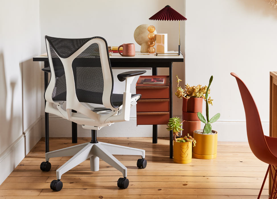 En blå och vit Cosm-kontorsstol från Herman Miller, placerad framför ett svart och terrakottafärgat Mode-skrivbord från Herman Miller. Placerat i ett hemmakontor med trägolv och vita väggar, kompletterat med en Matin-bordslampa och inomhusväxter.