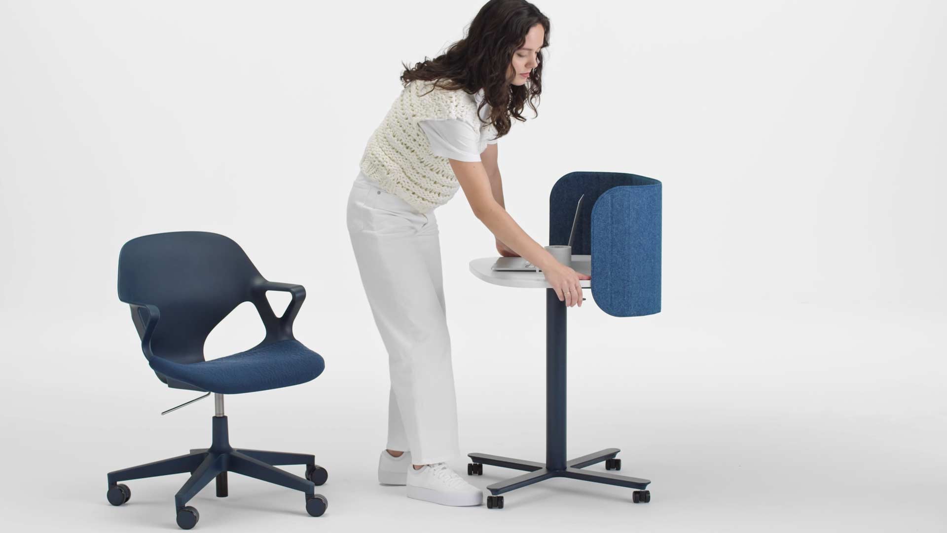 En video av en kvinna som sitter i en blå Zeph-kontorsstol och reser sig upp och höjer höjden på ett blått och vitt Passport-arbetsbord från Herman Miller med hjälp av spaken på sidan.