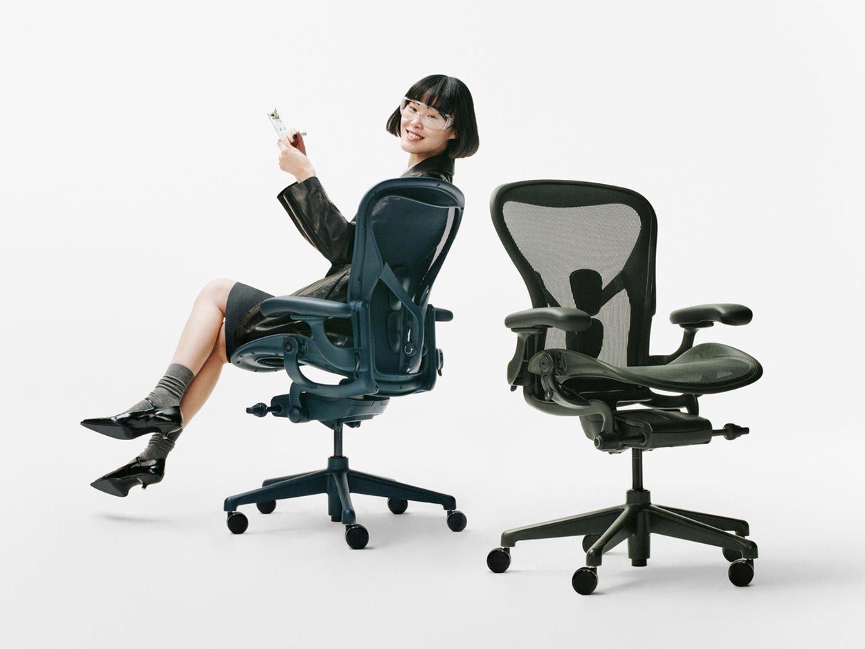 En kvinna satt på en blå Herman Miller Aeron-kontorsstol i färgen Nighfall bredvid en annan Aeron-stol i färgen Jasper Green i en vit ateljé.
