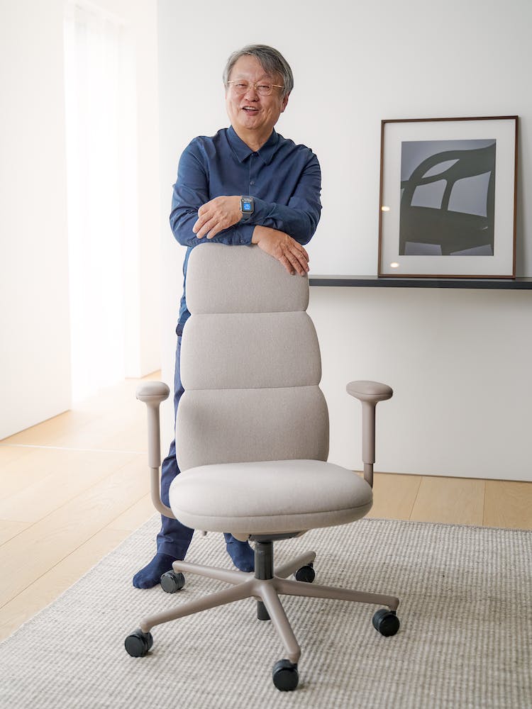 Designern Naoto Fukasawa lutad mot en Herman Miller Asari-kontorsstol. Miljön har ett rent golv och en ren vägg i bakgrunden.