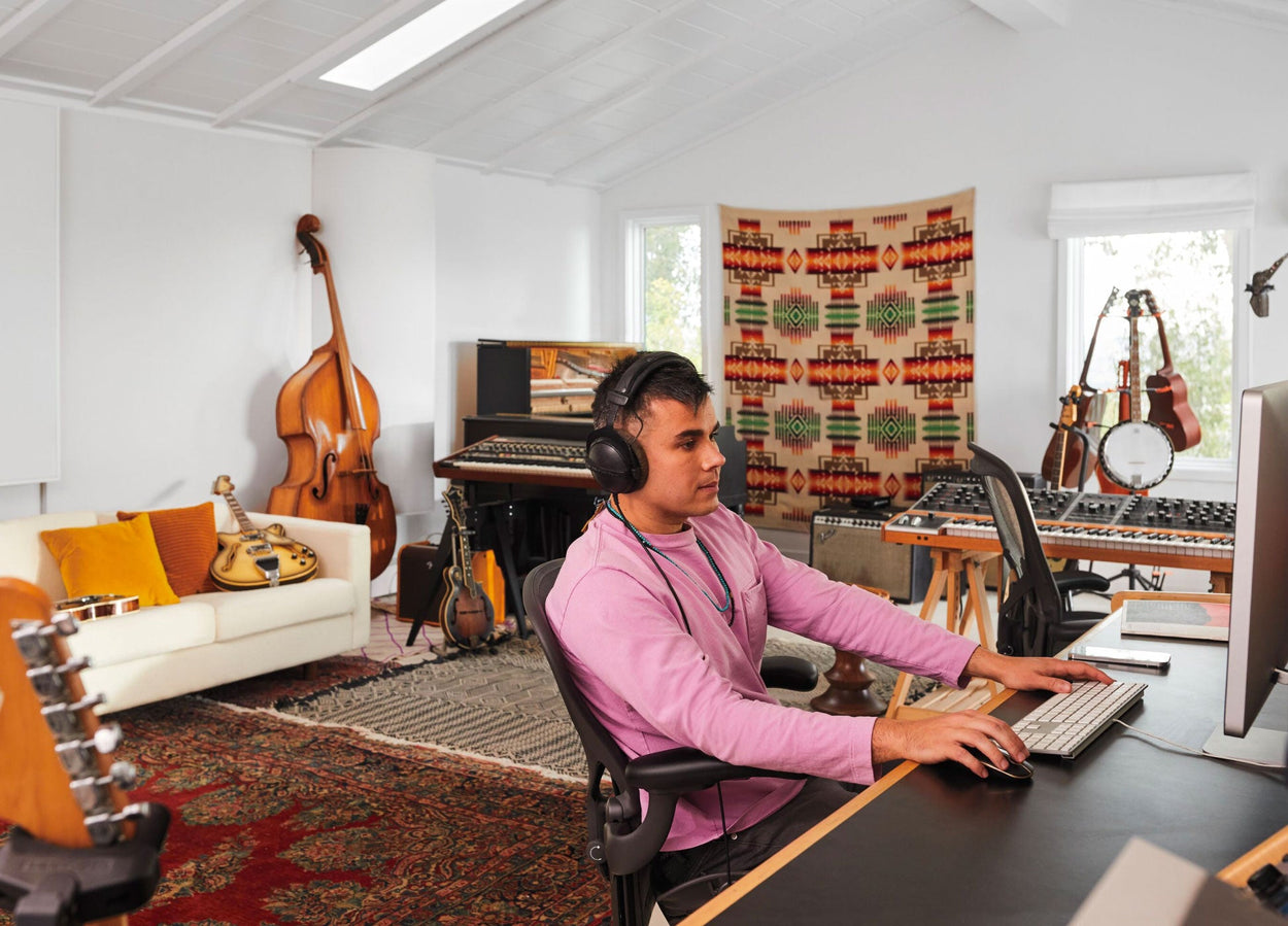 Rostam, musiker, producent och kompositör, sitter i en Aeron-kontorsstol i grafit vid en dator med hörlurar på och musikinstrument i bakgrunden.
