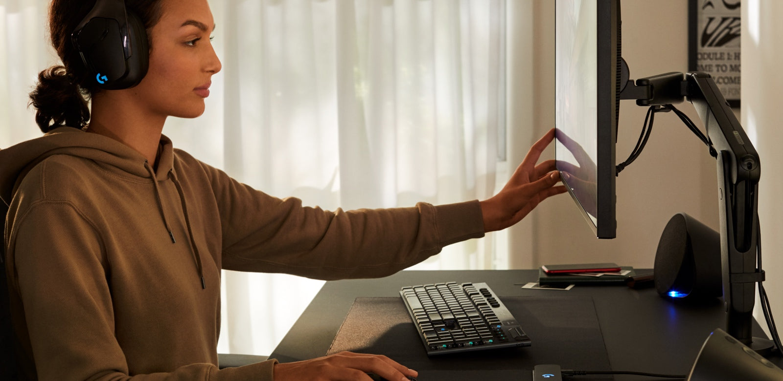 En kvinna sitter vid ett svart Nevi-spelbord med en svart Ollin-spelmonitorarm från Herman Miller. Hon har Logitech-hörlurar på sig och flyttar datorskärmen med ena handen.