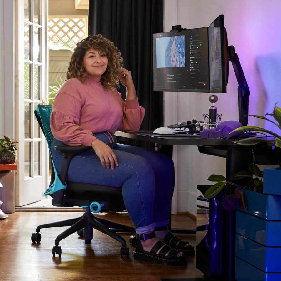 Proffsstreamer och röstskådespelare Friskk sitter på Herman Miller Sayl-gamingstol i oceanblått och svart, vid ett svart Nevi sit-stand-gamingbord med monitor på Oliin-monitorarm.