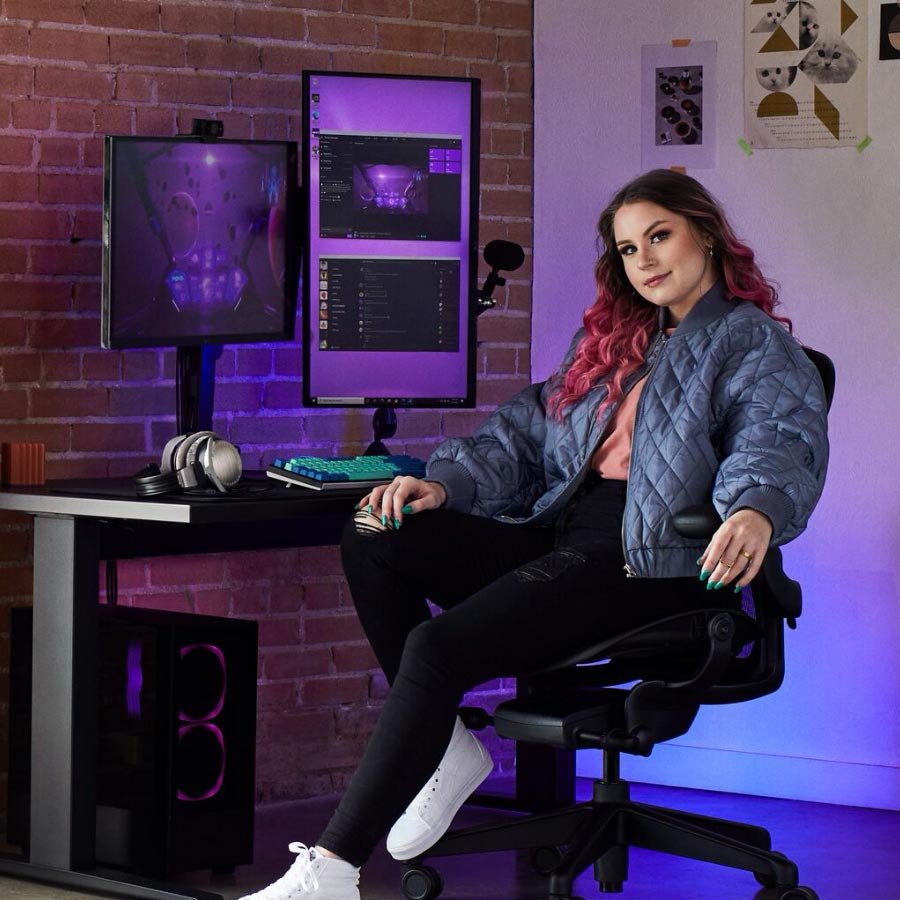 Stjärngamern och coachen Electra sitter vid en gaming-set med Herman Miller Aeron gamingstol och Nevi sit-stand-skrivbord i svart med dubbla skärmar på Ollin-skärmarmar