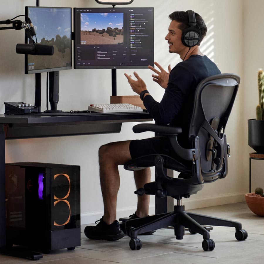 Gamern och streamern Xaryu i en gaming-uppställning sittande på Herman Miller Aeron gamingstol med svart Nevi sit-stand gamingbord och dubbla skärmar på Ollin-skärmarmar
