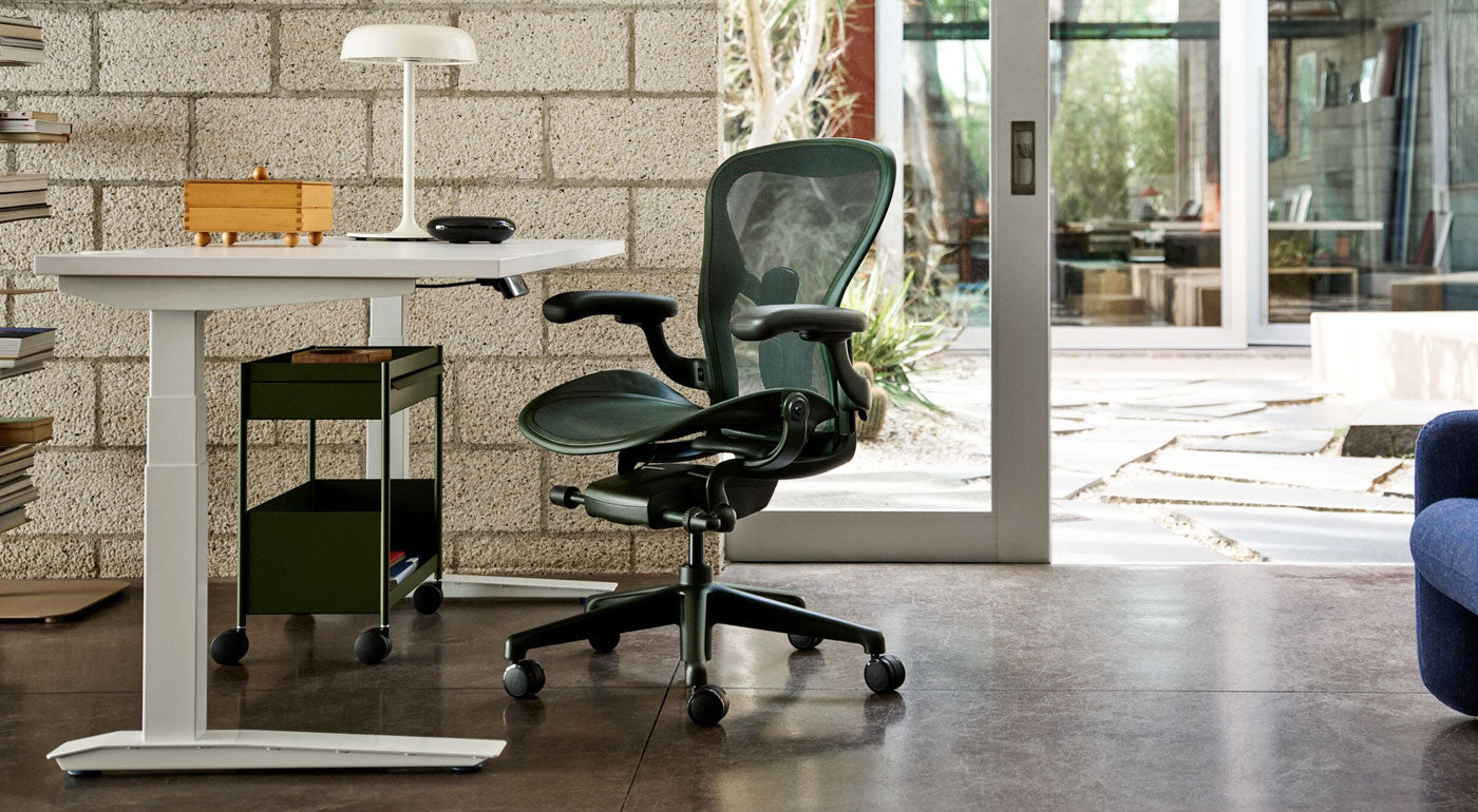 En Herman Miller Aeron-kontorsstol i färgen Jasper grön som en del av ett ljust hemmakontor med ett vitt Jarvis-skrivbord med höjdjustering och en olivgrön OE1-förvaringsvagn.
