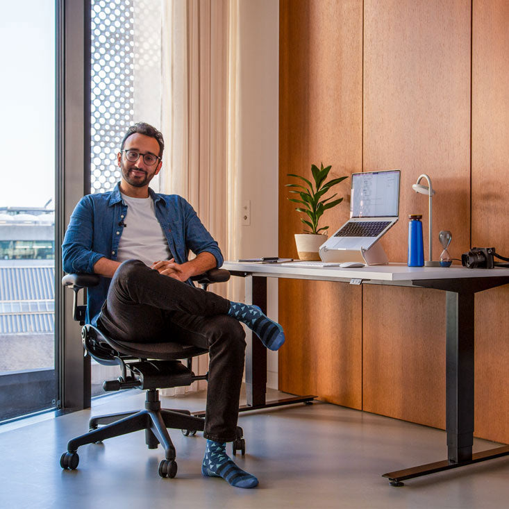Foto av Ali Abdaal, poddare, sittande i en Herman Miller Aeron kontorsstol i grafit framför ett Nevi sit-stand-skrivbord med svarta ben och vit skiva med en bärbar dator i ett Oripura-datorstativ