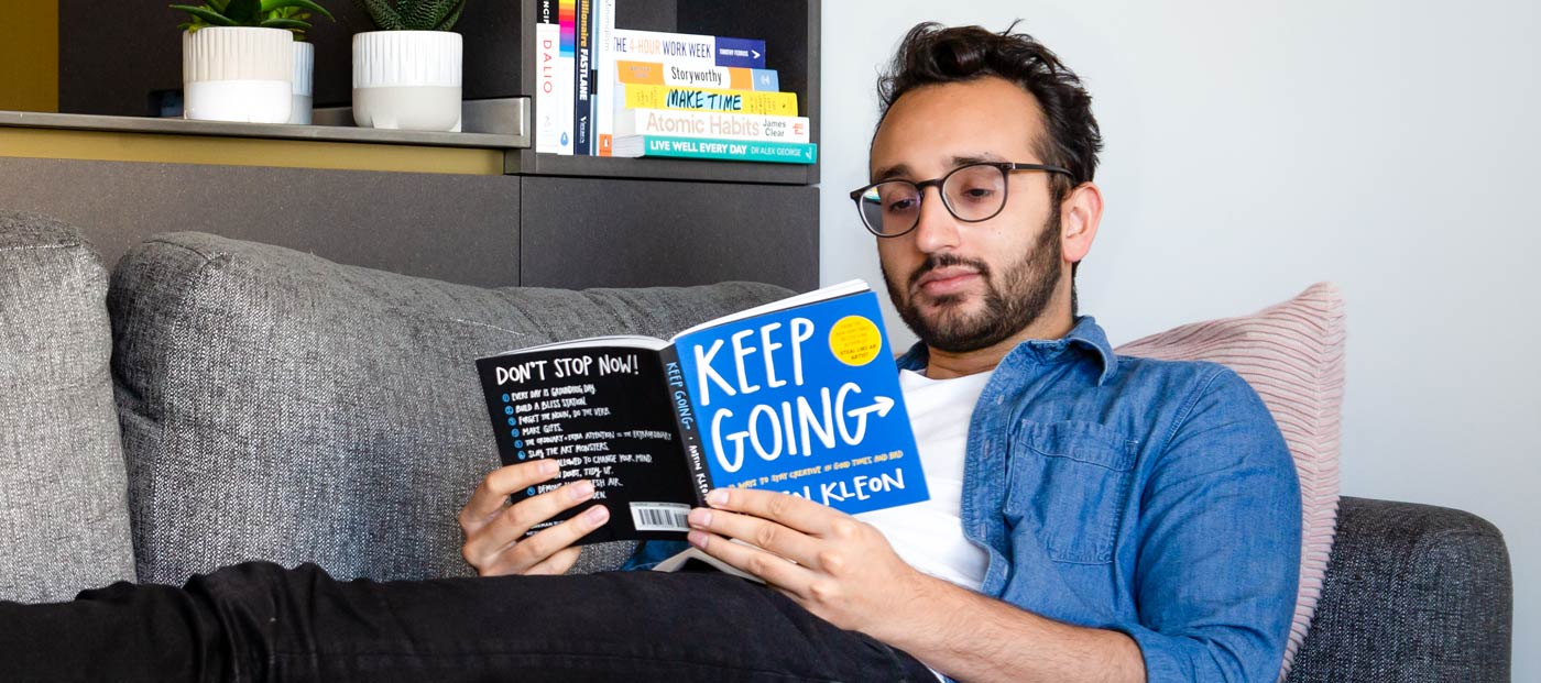 Foto av Ali Abdaal liggande på soffan och läsande en bok med titeln Keep Going