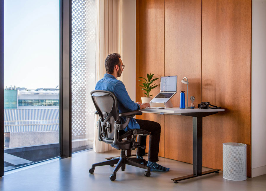 Foto av Ali Abdaal, poddare, sittande i en Herman Miller Aeron-kontorsstol i grafit framför ett Nevi-skrivbord med svarta ben och vit skiva och en bärbar dator i ett Oripura-laptopstativ