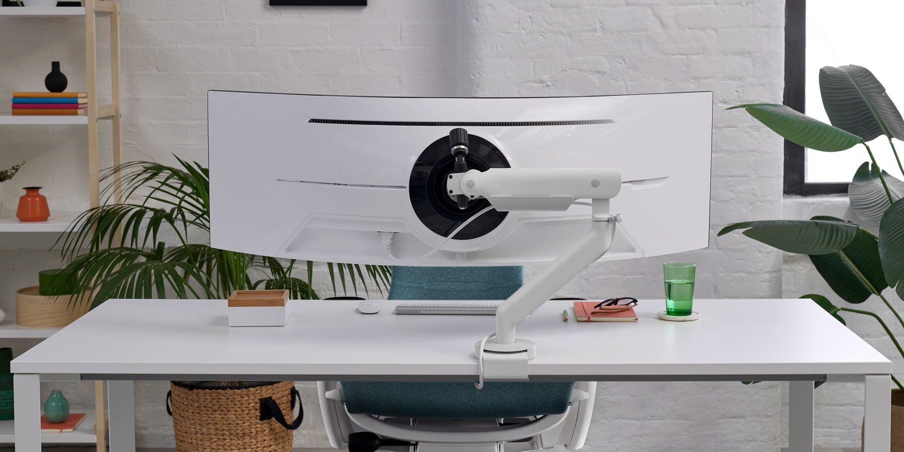 Framifrånvy av en vit Flo X-monitorarm från CBS fäst vid ett vitt skrivbord, placerad framför en svart Aeron-kontorsstol från Herman Miller.