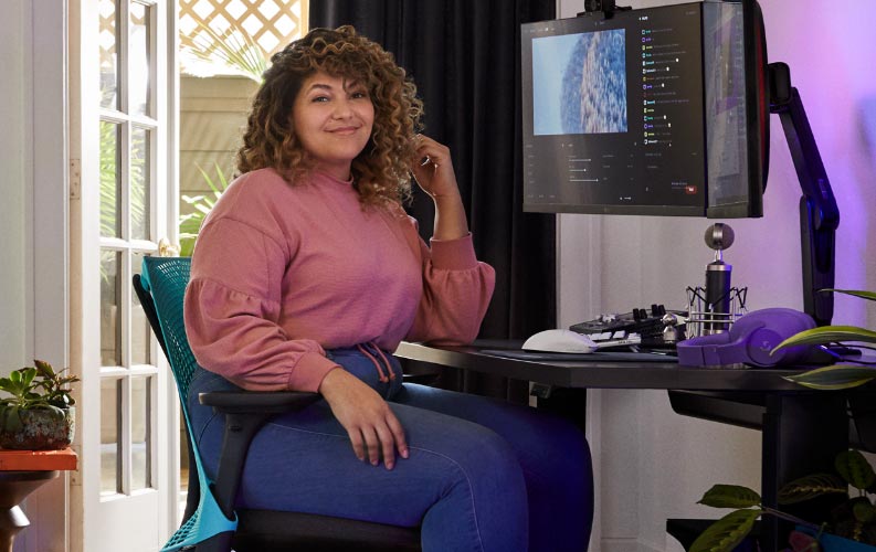 Proffsig streamer och röstskådespelare Friskk sitter på Herman Miller Sayl-gamingstol i djupblått och svart, vid ett svart Nevi-sit-stand-gamingbord med skärm på Oliin-monitorarm.