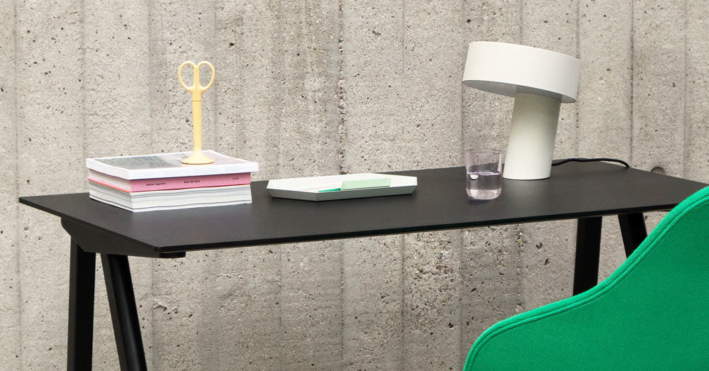 En vit Slant-lampa från HAY, placerad på ett svart Copenhague 90-skrivbord från HAY, bredvid en stapel böcker, gula saxar och ett vattenglas.