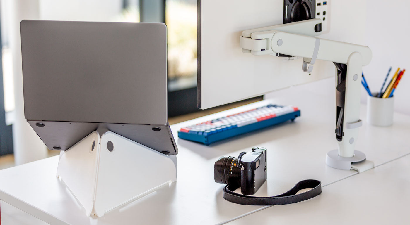 Ett vitt Oripura-laptopstativ från Herman Miller som håller en laptop bredvid en vit monitorarm.