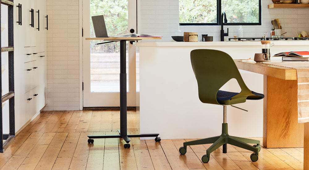 Ett svart och ekfärgat Passport-bord från Herman Miller placerat nära en vit köksö, med en bärbar dator och en bok. Bilden visar också en grön och svart Zeph-stol från Herman Miller placerad vid ett matbord i trä.