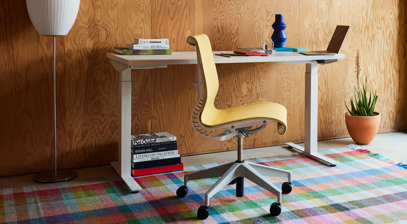En gul Setu-kontorsstol från Herman Miller, klädd i Mimosa Meld-tyg, placerad framför ett vitt Nevi-ståbord på en färgglad rutig matta.