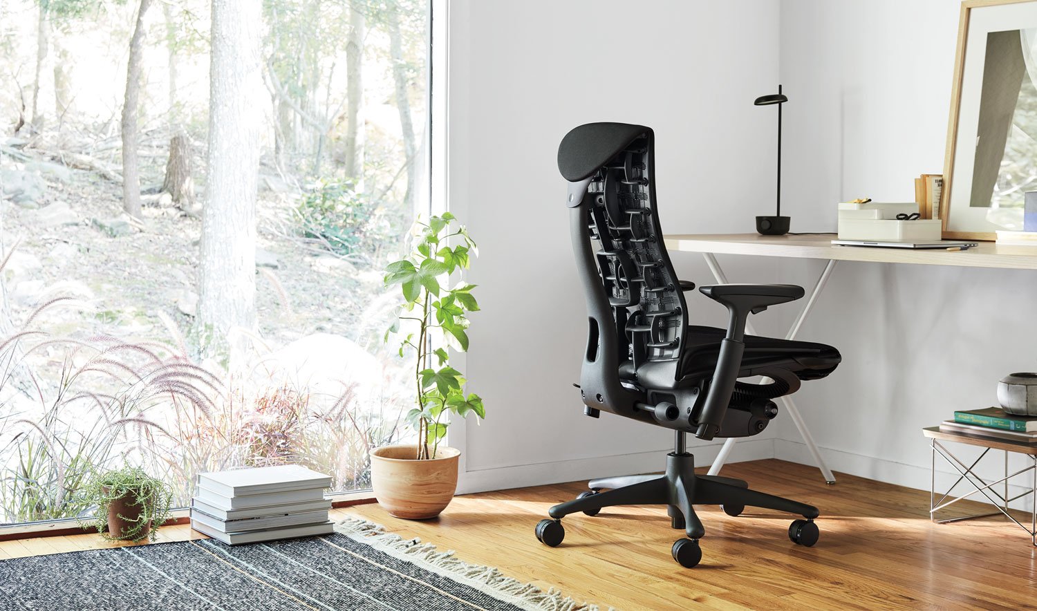 En svart Embody-kontorsstol från Herman Miller, placerad i en inomhusmiljö. Utrymmet innehåller element som möbler, golv och dekorativa föremål som en blomkruka och en vas.