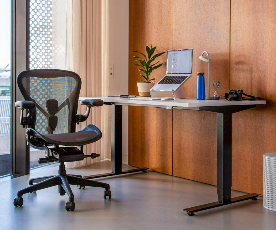 Hemmakontor med Nevi sit-stand-skrivbord i sittläge med svarta ben och vit skiva samt Aeron kontorsstol i grafit.