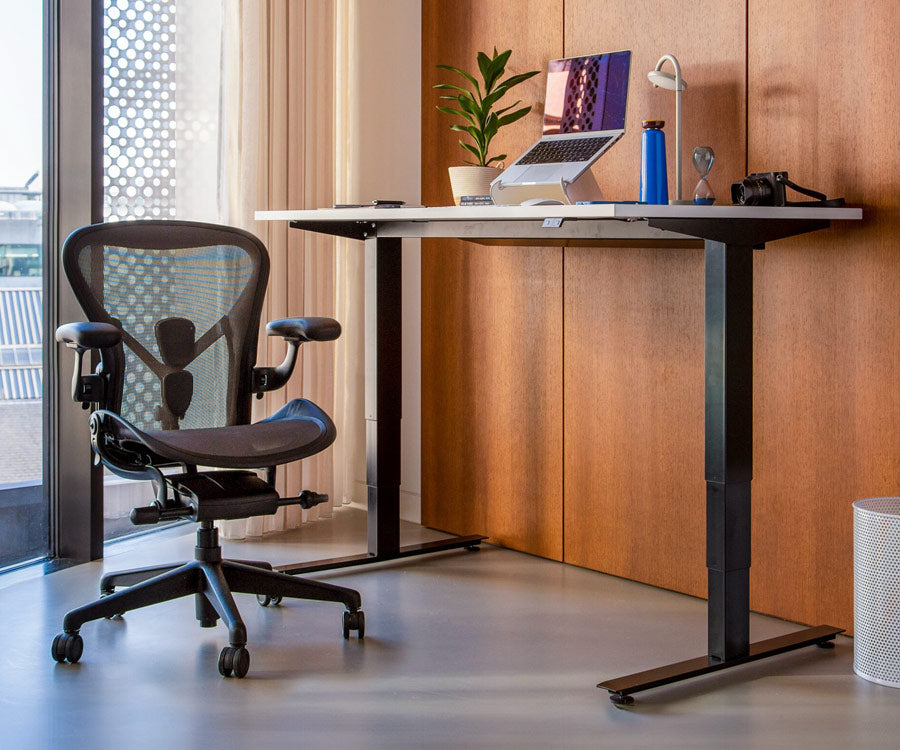 Hemmakontor med Nevi sit-stand-skrivbord i stående läge med svarta ben och vit skiva bredvid en Aeron-kontorsstol i grafit i bakgrunden.