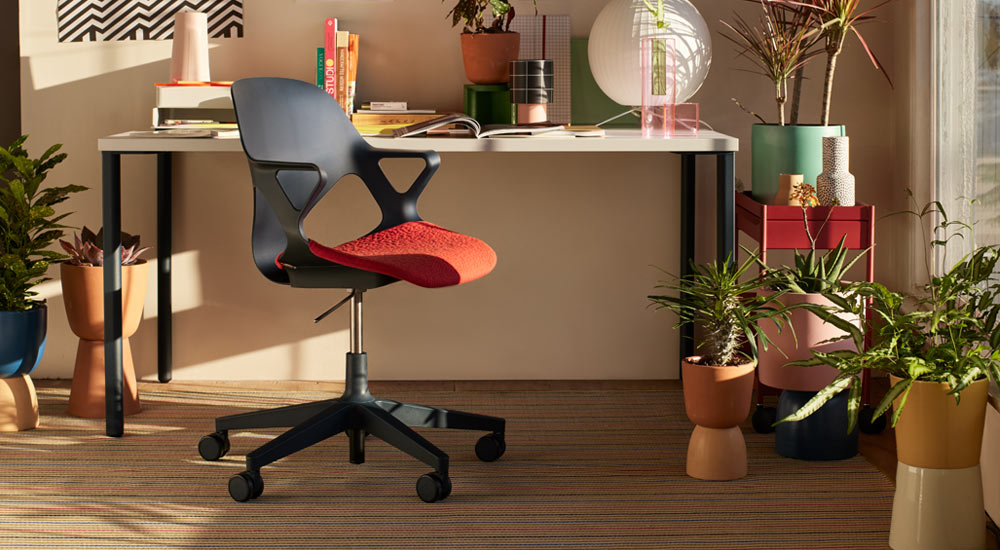 En svart Zeph-kontorsstol med röd sittdyna placerad framför ett Herman Miller OE1-skrivbord. Placerad i en hemmakontorsmiljö, med en röd OE1-vagn och ett urval av inomhusväxter i färgglada krukor.
