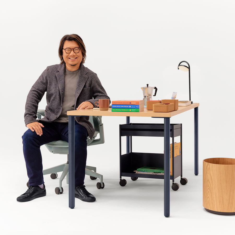 Bild av Kevin Kwan sittande i kontorsstolen Glacier Cosm med låg rygg i olika vinklar framför ett skrivbord OE1 med fast höjd i färgerna nightfall blue och ash med en förvaringsvagn OE1 under.
