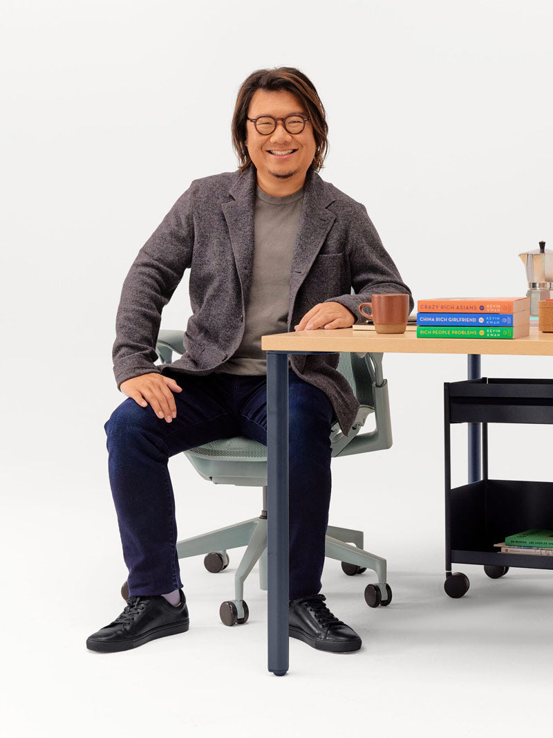 Glad bild av Kevin Kwan sittande i en Glacier Grey Cosm-kontorsstol lutad mot ett OE1-skrivbord med fast höjd, ben i Nightfall och ljus askfärgad skiva