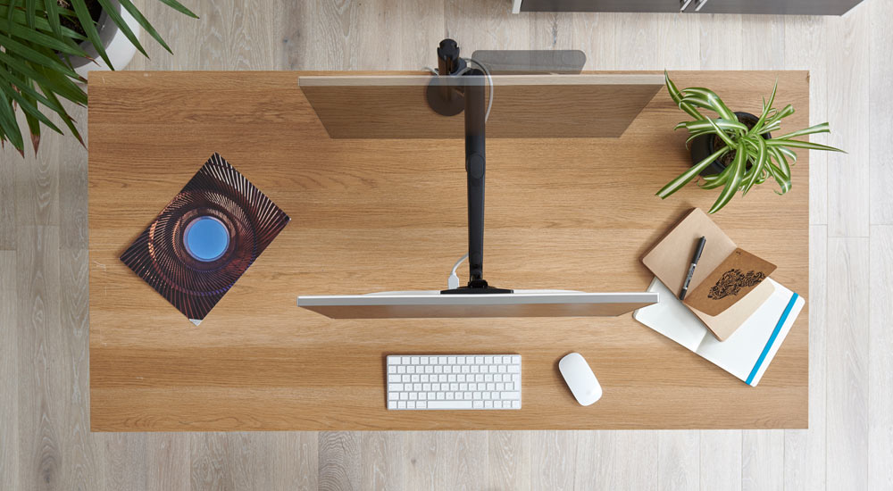 Ovanifrånvy av en svart Lima-monitorarm från CBS fäst vid ett träskrivbord, som visar monitorarmens och skärmens rörelse.