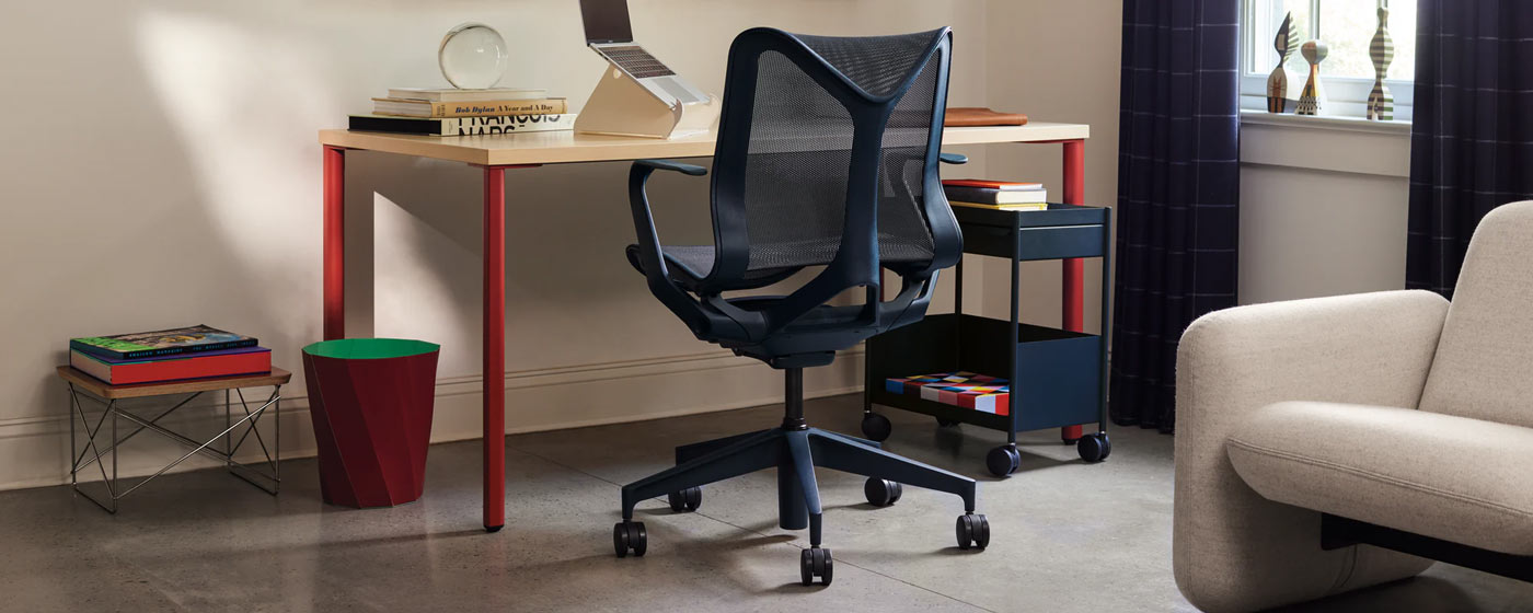 En blå Cosm-kontorsstol från Herman Miller, placerad framför ett rött och askfärgat OE1-skrivbord från Herman Miller. Placerad i ett hemmakontor, kompletterad med en blå OE1-vagn, ett vitt Oripura-laptopstativ och en röd HAY Paper-papperskorg.