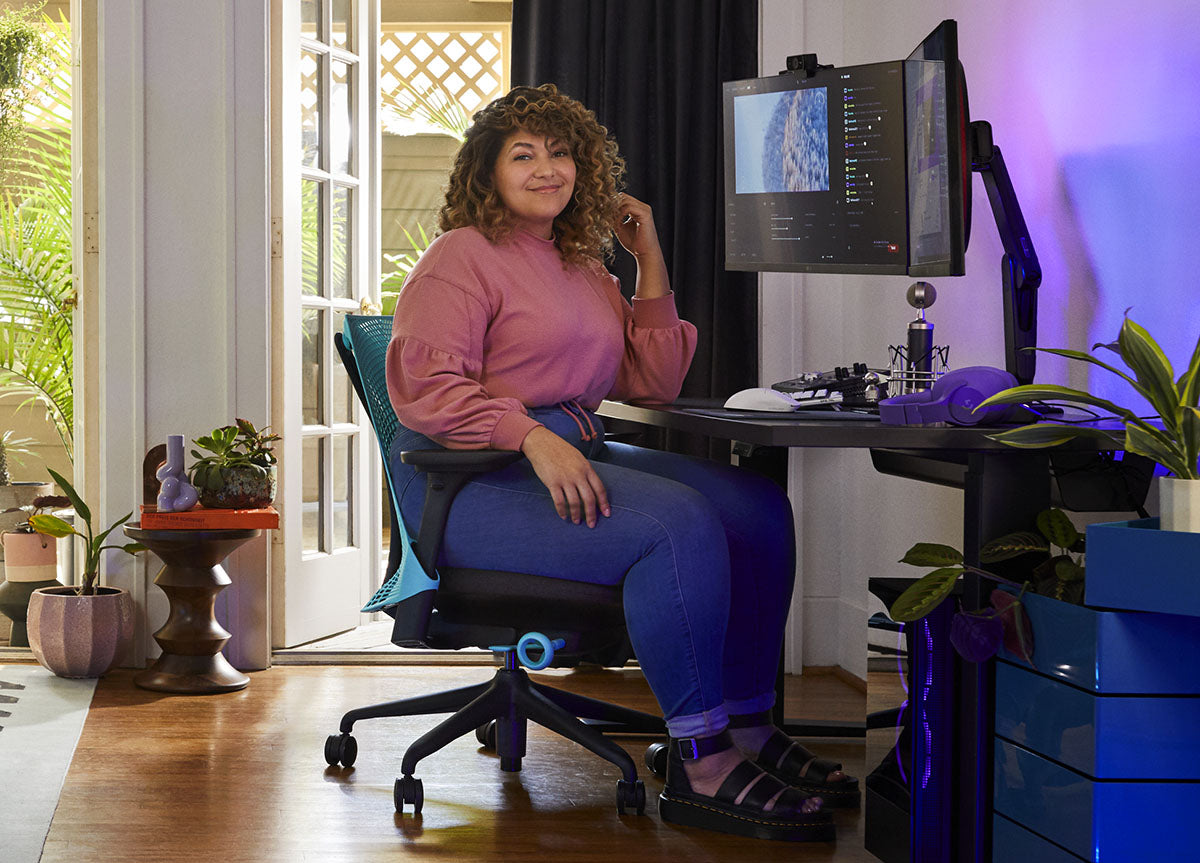 En kvinna sitter i en blå och svart Sayl-gamingstol från Herman Miller, placerad bakom ett svart Nevi-skrivbord med en svart Ollin-monitorarm och skärm. Miljön är en inomhusmiljö med trägolv, vita väggar och inomhusväxter.