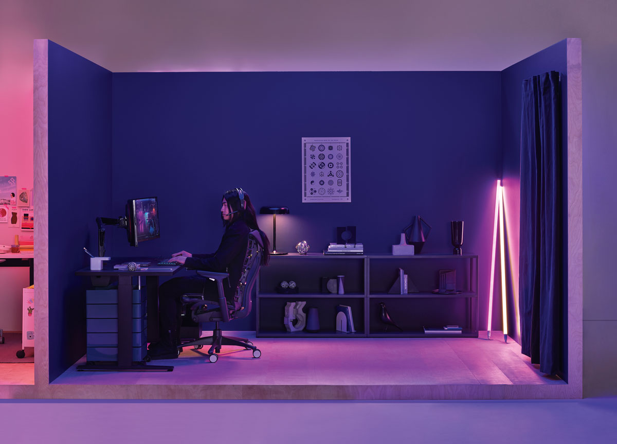 Spelkontor med kvinna som spelar sittande i en Logitech Embody spelstol i svart med ett Nevi spelbord i svart och Hay neonrörsbelysning i hörnet
