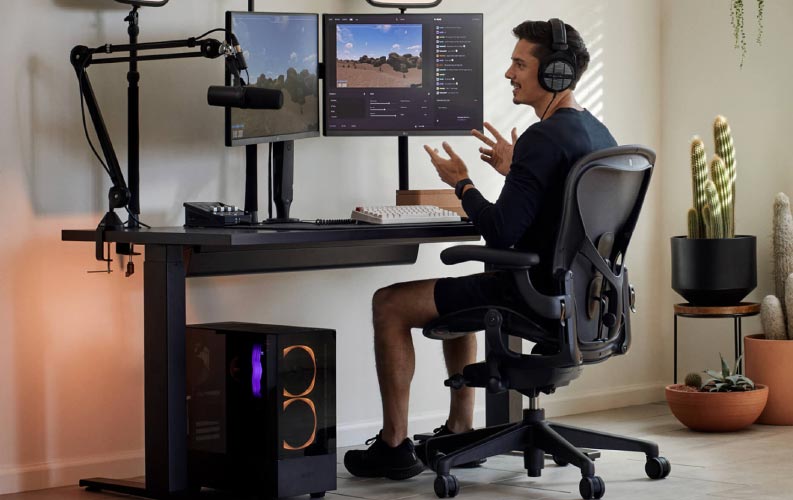Gamern och streamern Xaryu i en gaming-uppställning sittande på Herman Miller Aeron gamingstol med svart Nevi sit-stand gamingbord och dubbla skärmar på Ollin-monitorarmar