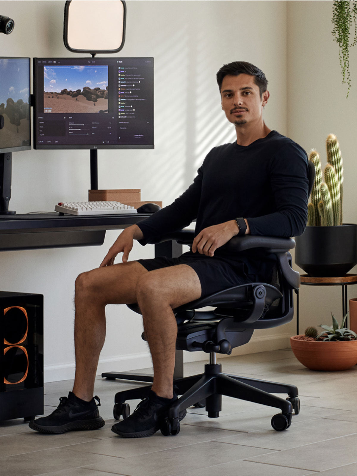 En man sitter i en svart Aeron-spelstol från Herman Miller, placerad bakom ett svart Nevi-skrivbord med en svart Ollin-monitorarm och skärm. Miljön är en inomhusmiljö med klinkergolv, vita väggar och krukväxter i terrakottakrukor.