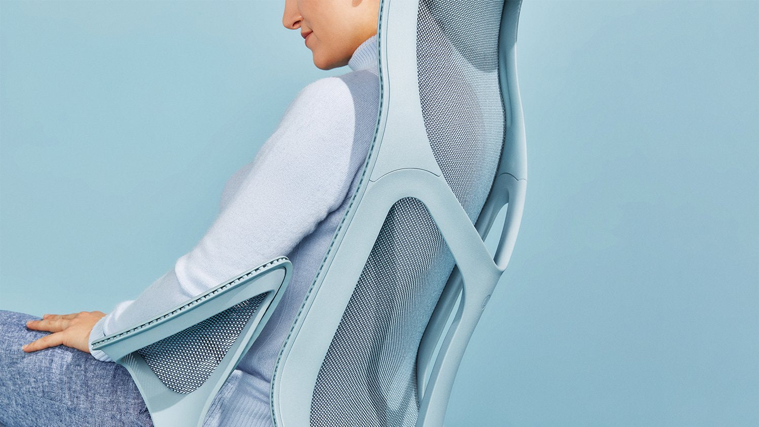 En person sitter i en mineralblå Cosm-kontorsstol från Herman Miller, klädd i en blå tröja och jeans framför en enfärgad ljusblå bakgrund.