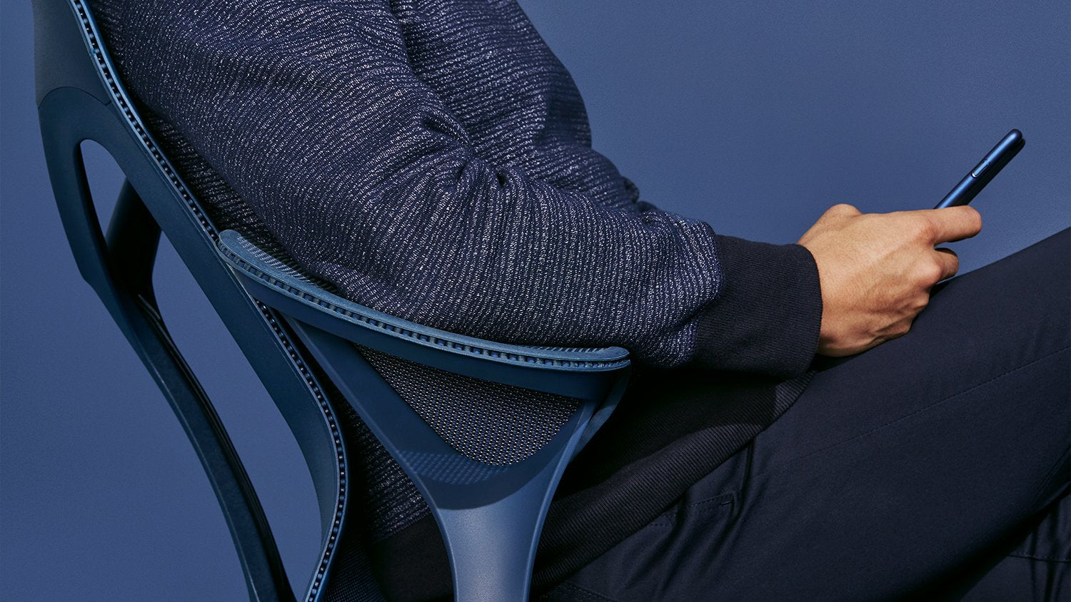 En person sitter i en nattblå Cosm-kontorsstol från Herman Miller. Personen håller en telefon och är klädd i mörkblå kläder framför en enfärgad mörkblå bakgrund.