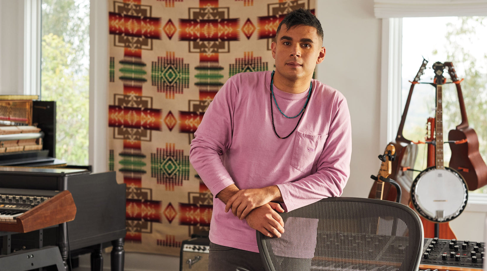 Rostam, musiker, producent och kompositör, står lutad mot en Aeron-kontorsstol i grafit med musikinstrument i bakgrunden.
